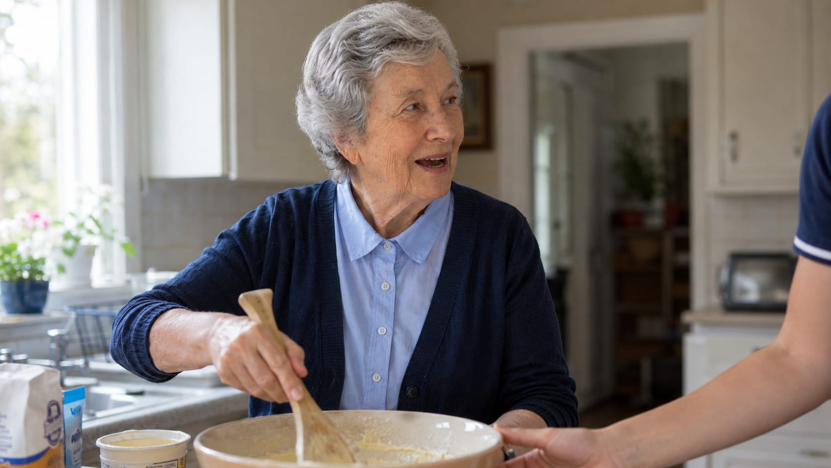 An older woman beats a mixture in a large bowl at her kitchen worktop, steadied by a hand at the rim.