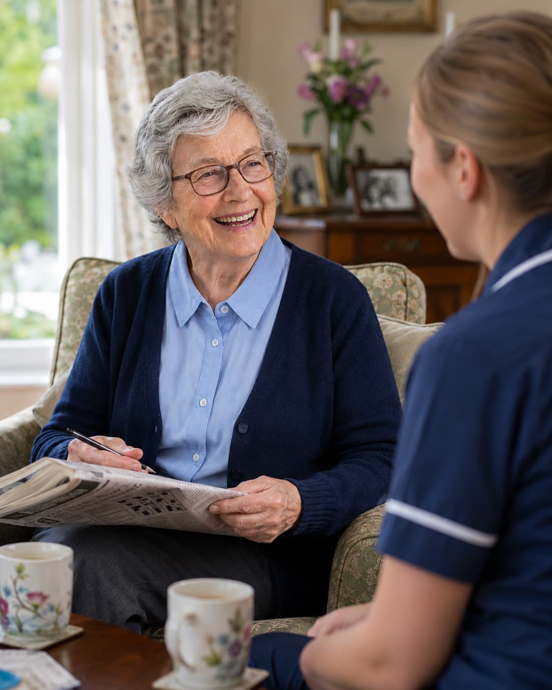An older woman in an armchair looks up from a folded newspaper, pen in hand, mid-sentence.