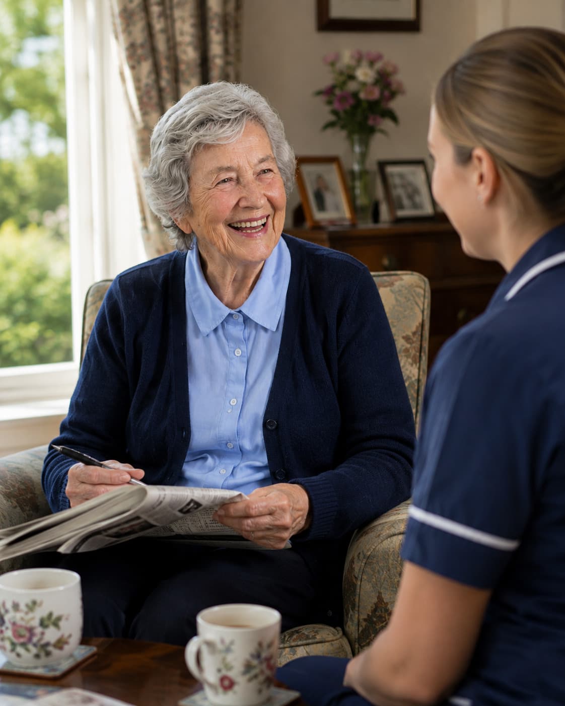 An older woman writes an answer into a crossword, laughing, a carer sitting opposite with two mugs between them.