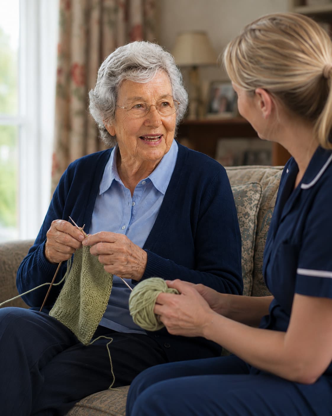 An older woman knits on a sofa by a window, talking to a carer sitting at the other end.
