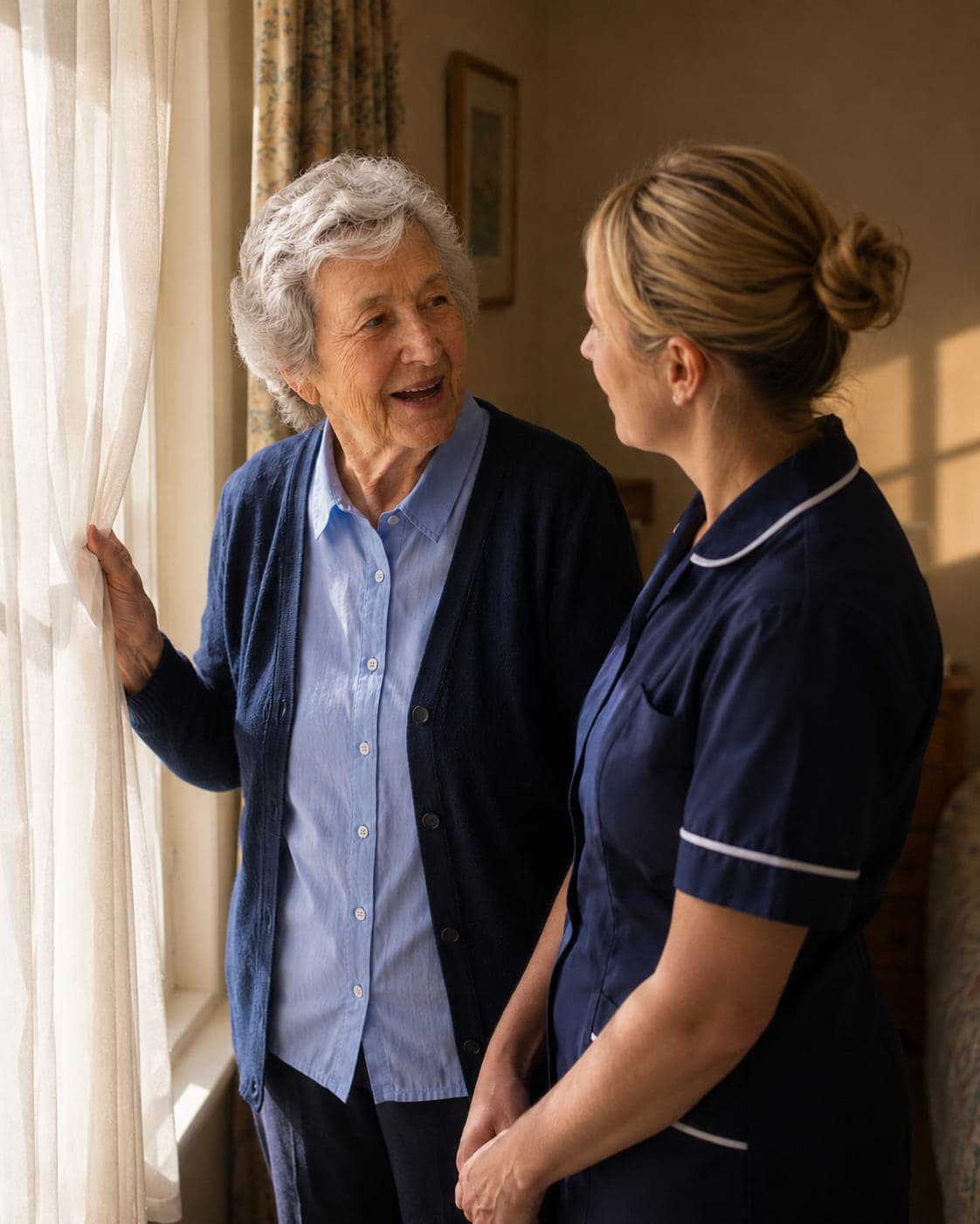 An older woman draws back her curtain to the morning, laughing at something the carer beside her has said.